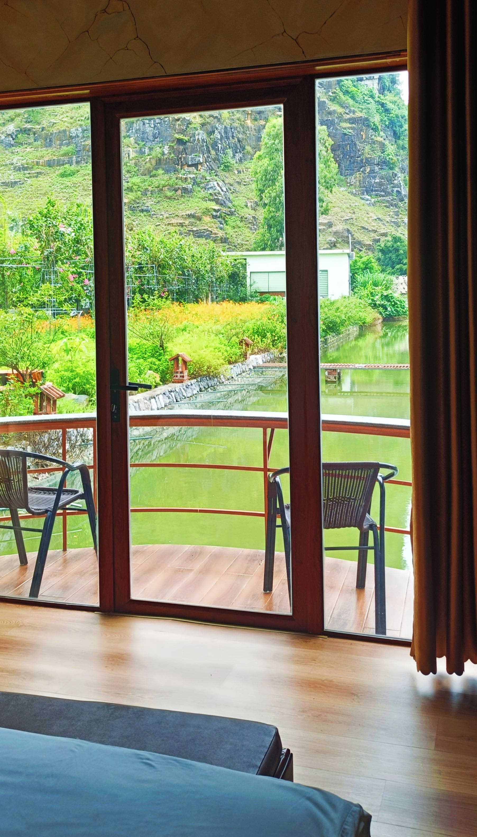 A room with a large window overlooking a garden and a pond. There are two chairs and a bed visible.