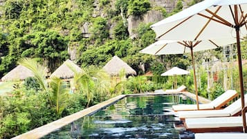 Un espace piscine agrémenté de chaises longues et de parasols, entouré d’une végétation luxuriante et de petits abris aux toits de chaume.