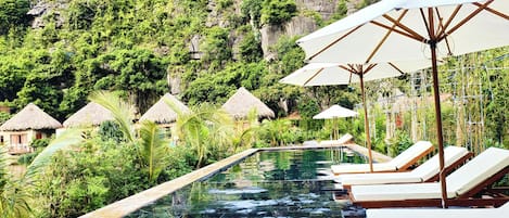 A pool area with lounge chairs and umbrellas, surrounded by lush greenery and thatched-roof huts.