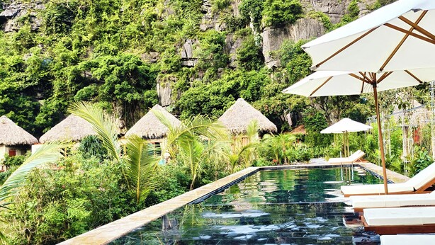 A pool area with lounge chairs and umbrellas, surrounded by lush greenery and thatched-roof huts. - Hang Mua Eco Garden (Hoa Lu)