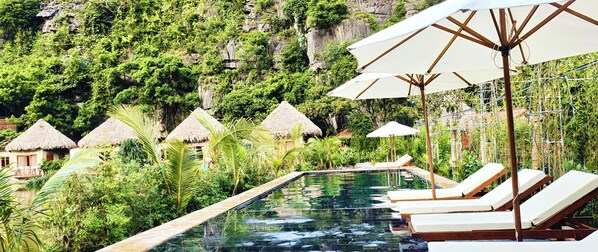 Un espace piscine agrémenté de chaises longues et de parasols, entouré d’une végétation luxuriante et de petits abris aux toits de chaume.