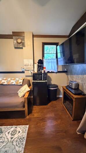 A compact living space with a sofa, a small kitchen area including a microwave, a toaster, and a coffee maker, a television mounted on the wall, and a window with a view of greenery. - Gray’s Riverside Camp Cabin 4 (Big Rapids)
