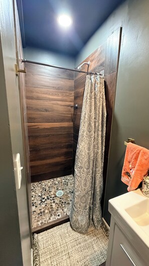 Une douche avec un panneau décoratif en bois, un rideau de douche à motifs et un sol carrelé avec un motif de galets.