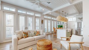 A spacious living room with a large sofa, a round wicker ottoman, and a wooden chair. A kitchen with a bar area is visible in the background. - Resort Style Pools, Golf Cart Boardwalk to the Beach (Port Aransas)
