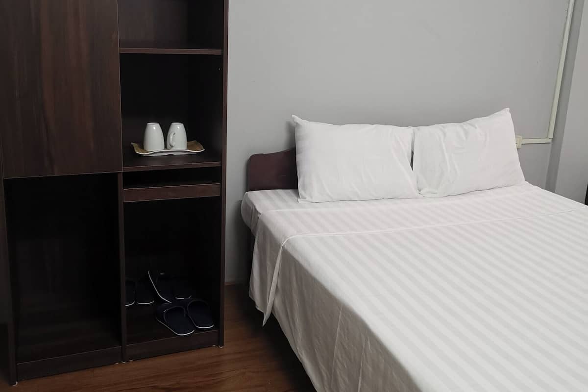 Quarto com cama, guarda-roupa e prateleira com chávenas e chinelos.