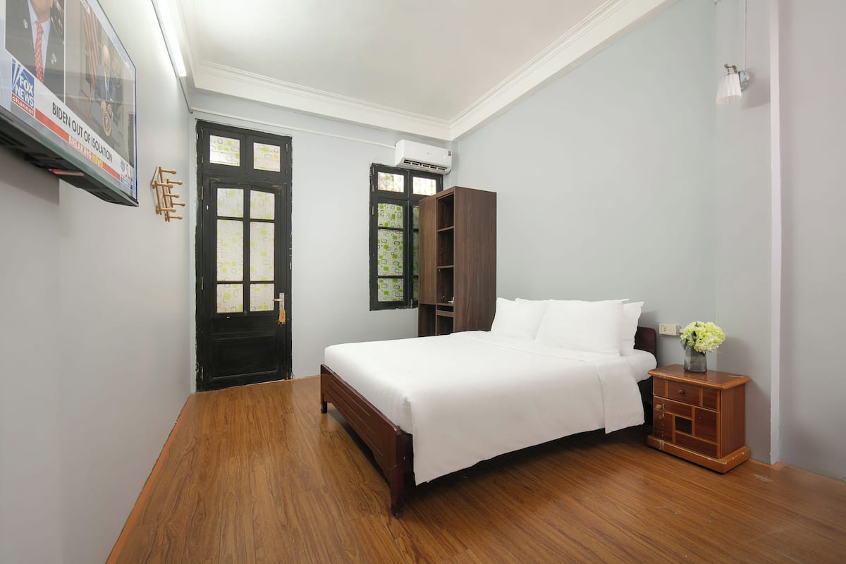 Quarto bem arrumado com piso de madeira, uma cama com roupa de cama branca, uma mesa de cabeceira de madeira e uma TV fixada na parede exibindo um canal de notícias.