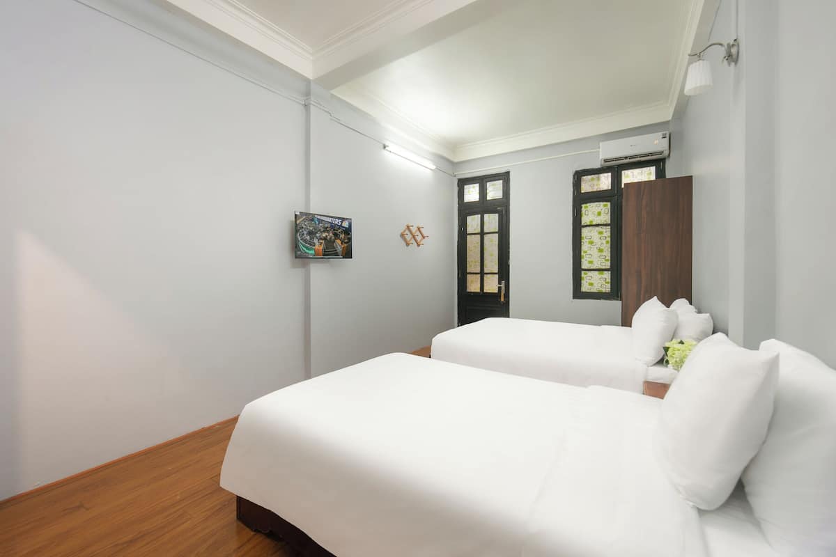 Quarto de hotel com duas camas, piso de madeira, parede branca, quadro emoldurado, porta com painéis de vidro e um aparelho de ar condicionado.