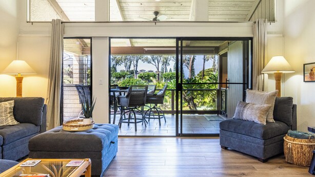 A spacious living area with two sofas, a coffee table, and a dining table with chairs. A sliding glass door leads to an outdoor patio with a view of greenery. - Breathtaking Condo - Ocean View - Now With AC! (Waikoloa)