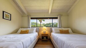 A room with two beds, a nightstand with a lamp, a framed picture on the wall, and a window with a view of trees. - Breathtaking Condo - Ocean View - Now With AC! (Waikoloa)