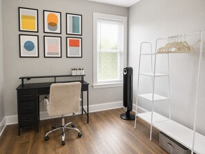 Ein Zimmer mit Schreibtisch, Stuhl, Fenster, Heizkörper und einem weißen Regal.