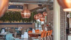 A modern restaurant interior with round tables, blue and orange chairs, and copper pendant lighting.