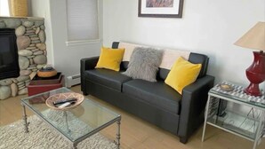 Een woonkamer met een zwarte bank, twee gele kussens, een glazen salontafel, een stenen openhaard, een ingelijste foto aan de muur, en een rode lamp op een bijzettafeltje.