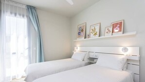 A hotel room with two beds, a white headboard, a bedside table, and a wall-mounted shelf with framed artwork. - Home2Book Stylish Apartment Pool & Terrace Abades (Abades)