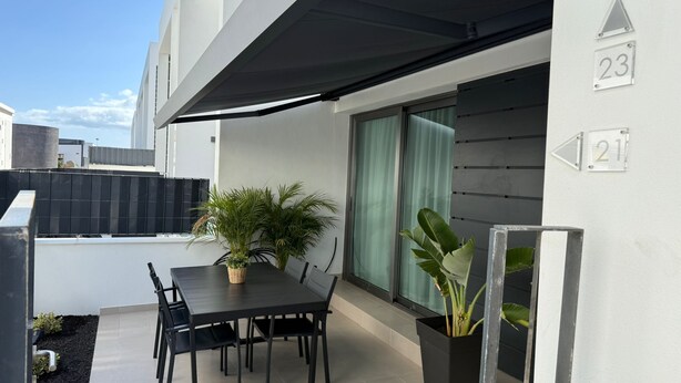 A modern outdoor dining area with a table and chairs, potted plants, and a large window with curtains. - Home2Book Stylish Apartment Pool & Terrace Abades (Abades)