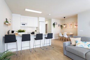 A modern kitchen with white cabinets, a microwave, and a coffee machine. There are four bar stools and a grey sofa in the living area. - Home2Book Stylish Apartment Pool & Terrace Abades (Abades)