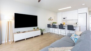 A modern living room with a grey sofa, a flat-screen TV, a ceiling fan, and a kitchen area with white cabinets and a dining set. - Home2Book Stylish Apartment Pool & Terrace Abades (Abades)
