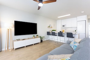 A modern living room with a grey sofa, a flat-screen TV, a ceiling fan, and a kitchen area with white cabinets and a dining set. - Home2Book Stylish Apartment Pool & Terrace Abades (Abades)