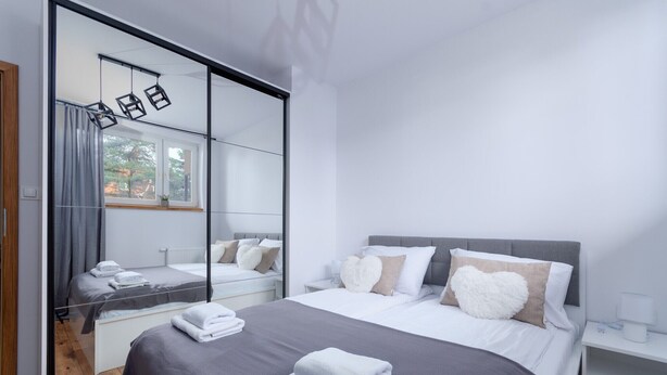 A modern bedroom with a large mirror, a bed with grey bedding and heart-shaped pillows, and a bedside table with a lamp. - Termy Cieplice Apartment (Jelenia Góra)