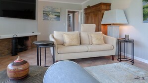 A living room with a sofa, two side tables, a flat-screen TV, a fireplace, and a painting on the wall. - Strand Cottage - Ferienwohnung "strand-cottage" in Westerland (Sylt)