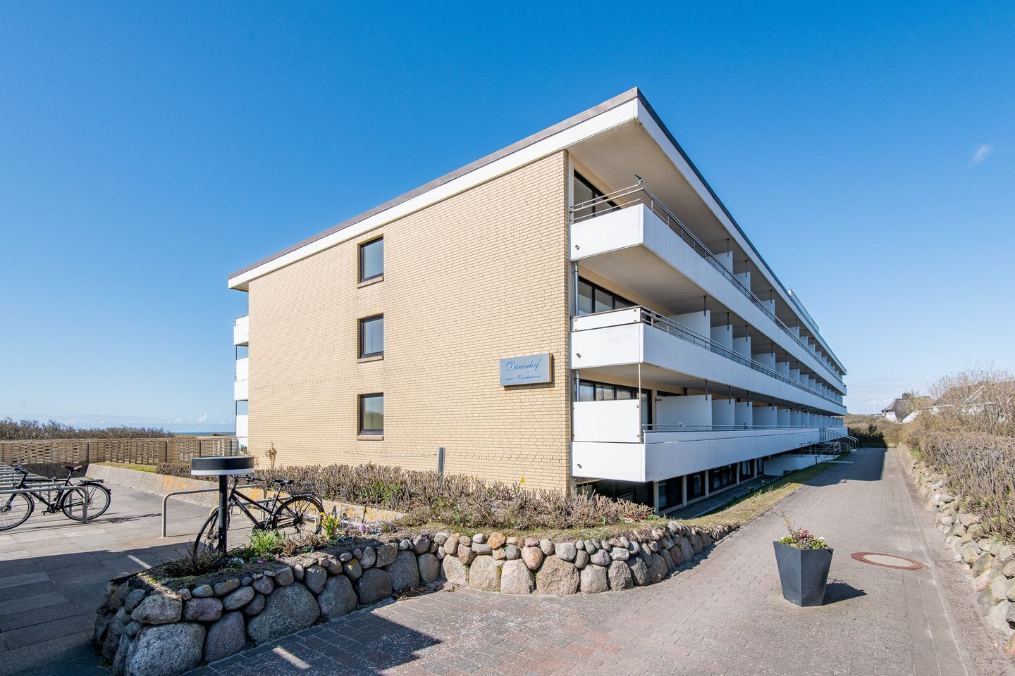 A modern multi-story building with a flat roof, a stone retaining wall, and a paved pathway leading to the entrance.