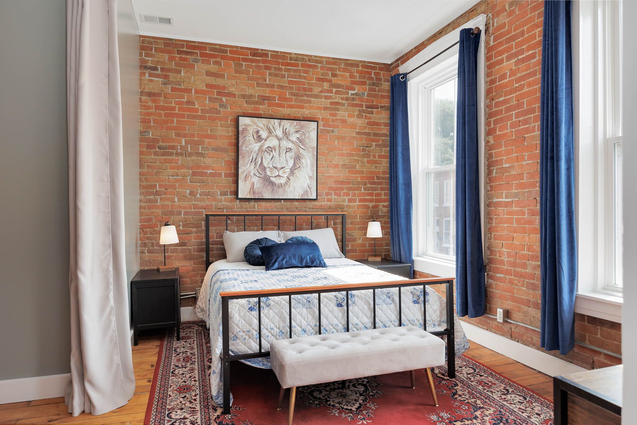 A bedroom with a brick wall, a bed with a headboard, a bench, a nightstand, a lamp, and a window with curtains.