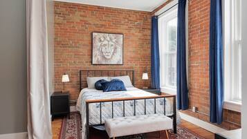 A bedroom with a brick wall, a bed with a headboard, a bench, a nightstand, a lamp, and a window with curtains.
