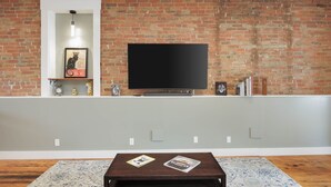 Un salón con pared de ladrillo, un televisor de pantalla plana montado sobre una estantería y una mesa de centro con revistas.