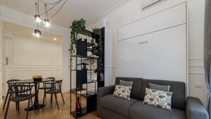 A living room with a grey sofa, black chairs, a black shelving unit with plants, and a dining table with chairs. - Toscana 2bis - FromHometoRome (Roma)