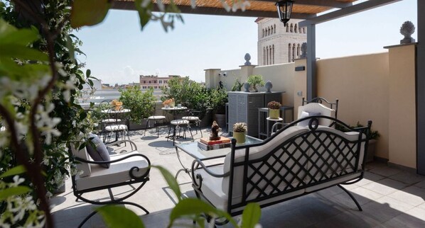 A rooftop patio with outdoor furniture, potted plants, and a view of buildings in the background.