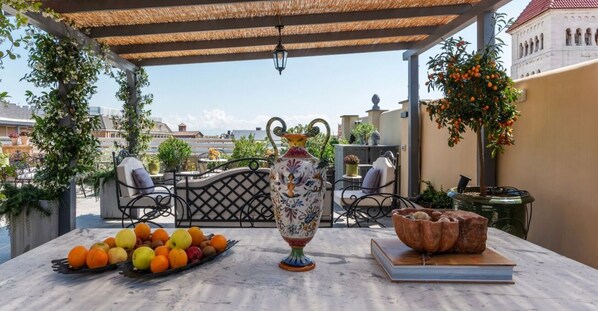 Une table avec des fruits, un vase décoratif et un livre, sur une terrasse offrant une vue sur les bâtiments.