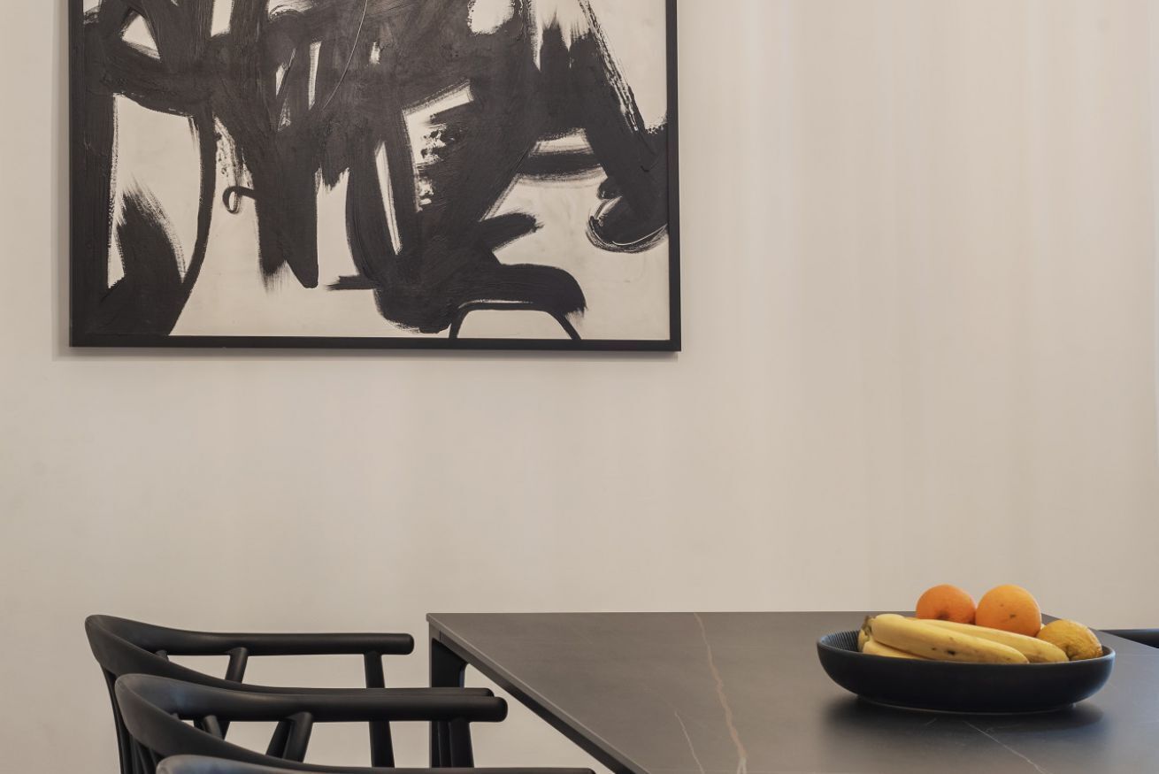 A dining area with a table and chairs, a black abstract painting on the wall, and a bowl of fruit on the table.