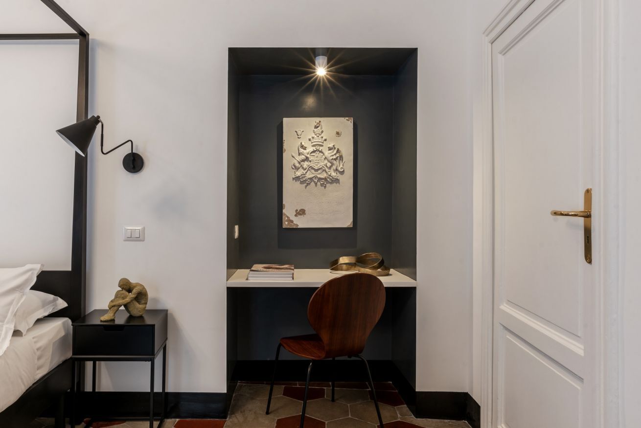 A bedroom with a bed, a desk with a chair, a wall-mounted lamp, a decorative piece, and a door.