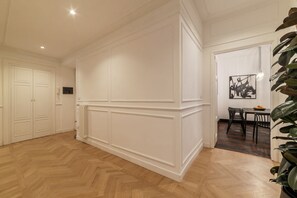 Ruime kamer met een houten parketvloer, witte lambrisering, een eethoek met tafel en stoelen, en een grote plant.