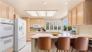 A modern kitchen with wooden cabinets, a large island with bar stools, built-in appliances, and a view of greenery outside.