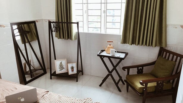 A room with a bed, a chair, a small table with a teapot and cup, a laptop on the bed, and a window with curtains. - Moonie House Saigon (Ho Chi Minh City)