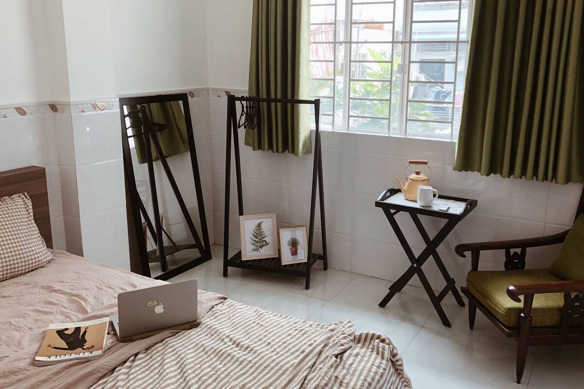 Ein Schlafzimmer mit einem Bett, einem Laptop, einem Stuhl, einem kleinen Tisch mit Teekanne und Tasse sowie einem Fenster mit Vorhängen.