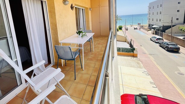 Uma varanda com mesa e cadeiras, vista para a rua e o mar, e um prédio com uma placa.