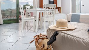 Une pièce moderne avec une grande fenêtre, une table à manger, des chaises blanches, un canapé et un panier en osier contenant un chapeau et une télécommande.