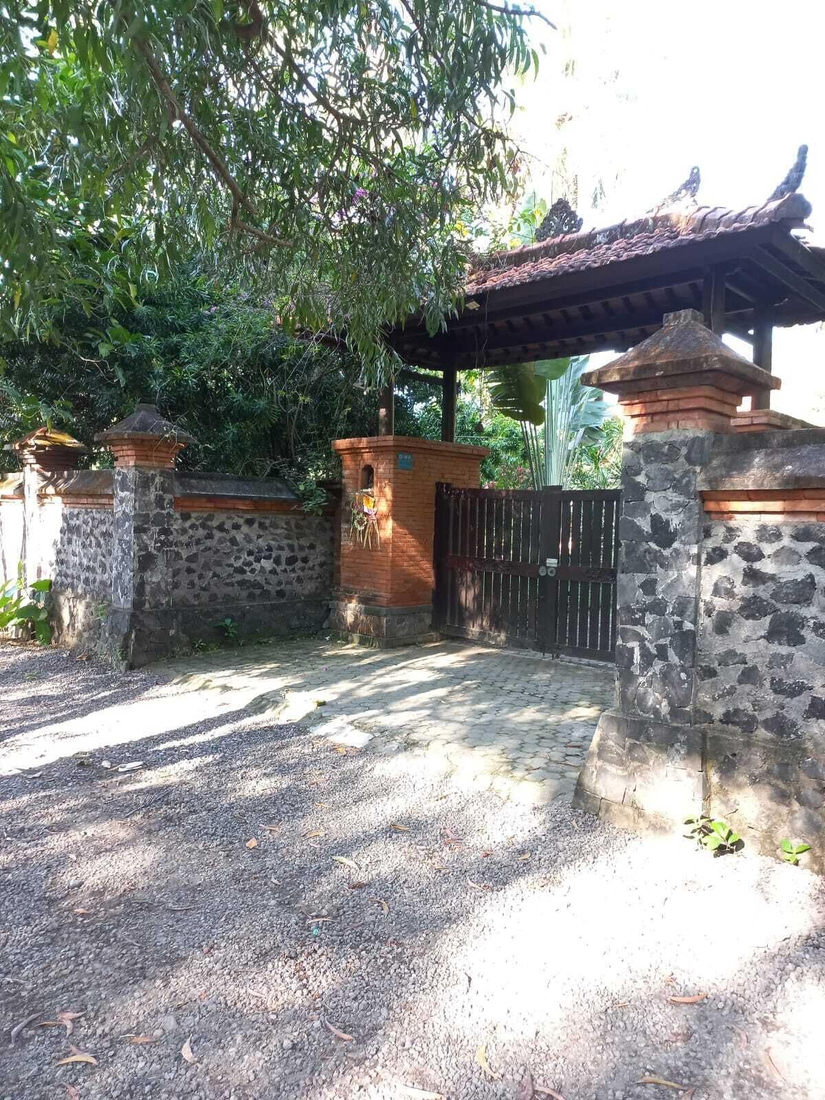 Uma entrada murada com paredes de pedra e portão de metal, cercada por vegetação e uma estrutura de telhado.
