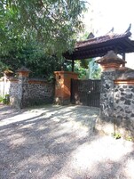 Uma entrada murada com paredes de pedra e portão de metal, cercada por vegetação e uma estrutura de telhado.