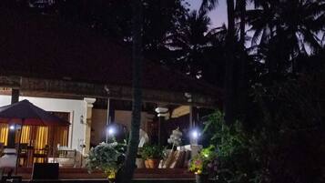 A nighttime view of a pool area with palm trees and a building with a covered seating area.