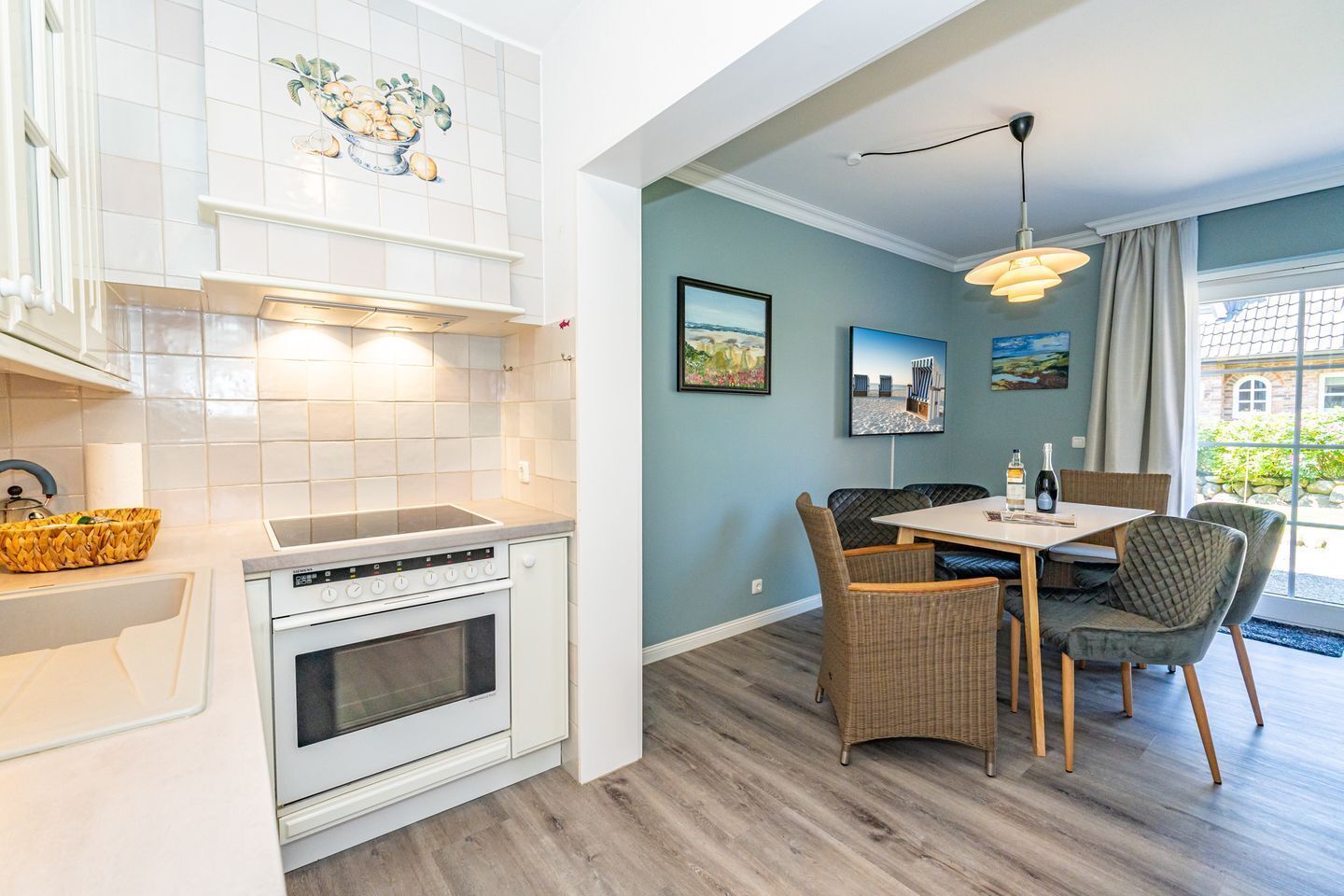 A modern kitchen with white cabinets, a built-in oven, and a dining area with wicker chairs and a table.