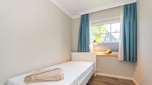 Ein kleines, ordentlich eingerichtetes Schlafzimmer mit einem Einzelbett, einem Fenster mit Vorhängen und einem kleinen Tisch mit einer Lampe.