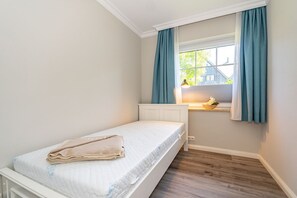 Une petite chambre à coucher, bien rangée, avec un lit simple, une fenêtre avec des rideaux et une petite table sur laquelle se trouve une lampe.