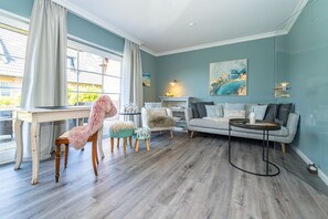 A living room with a sofa, a round coffee table, and a chair. There is a window with curtains, a painting on the wall, and a small table with a lamp. - Nis Puk - vacation apartment in Westerland for max. 3 persons, with west-facing terrace and (Sylt)
