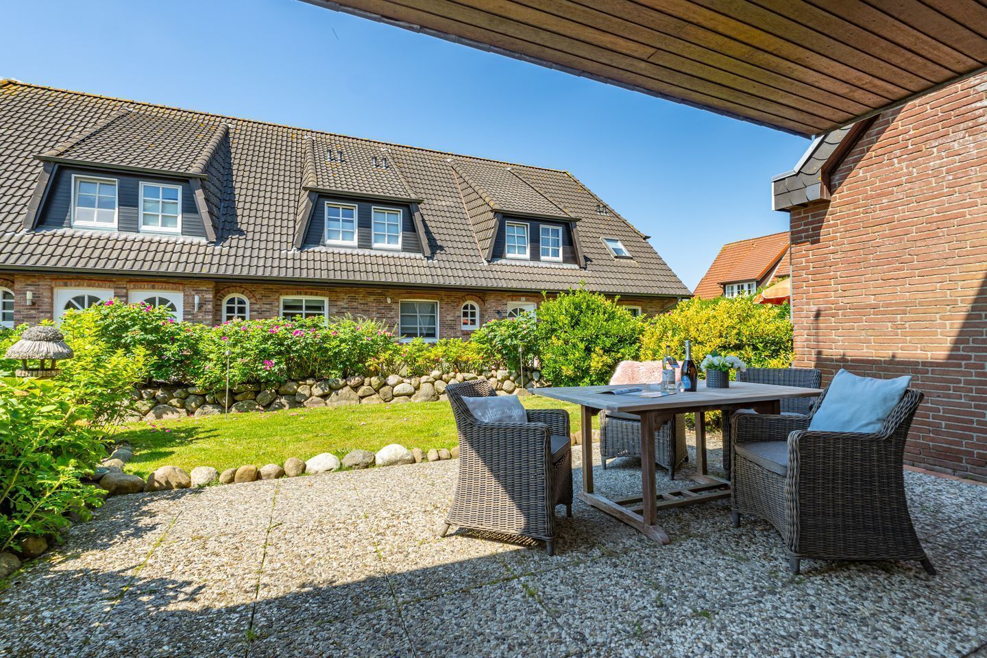 A house with a patio area featuring a table and wicker chairs, surrounded by a garden with a stone wall and greenery.