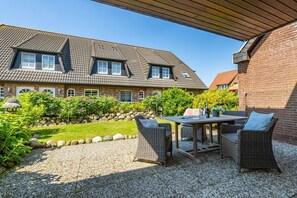 A house with a patio area featuring a table and wicker chairs, surrounded by a garden with a stone wall and greenery.