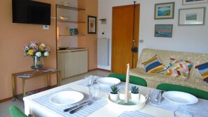 A dining area with a table set for four, a television mounted on the wall, and a sofa with colorful cushions.