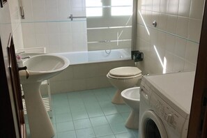 A bathroom with a sink, two toilets, a bathtub, and a washing machine.
