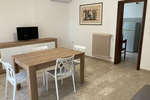 A dining area with a wooden table and white chairs, a television on a stand, a radiator, and a framed picture on the wall.
